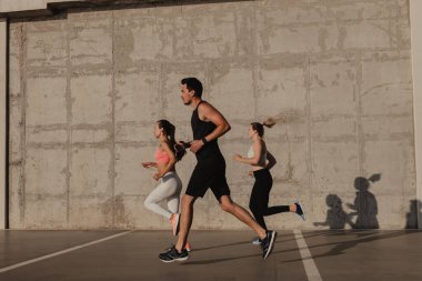 Spor kıyafetleri içinde, beton duvarın önünde birlikte koşan kendinden emin üç genç insan.