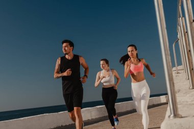 Spor giyimli üç kendine güvenen genç, birlikte deniz kenarında koşarken konsantre görünüyorlar.