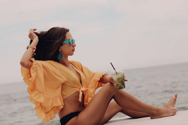 海辺をリラックスしながらカクテルを楽しむブーホースタイルの作物トップに魅力的な若い女性 — ストック写真