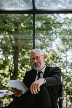Dijital tablet tutan ve ofiste otururken gülümseyen kendinden emin bir iş adamı.