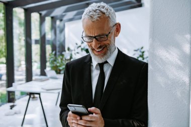 Akıllı telefon kullanan ve modern ofiste dururken gülümseyen kendinden emin bir iş adamı.