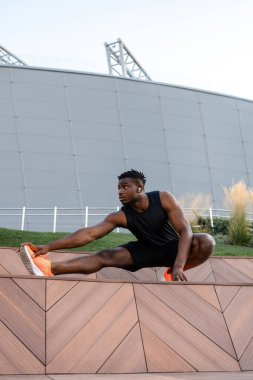 Spor kıyafetleri içinde konsantre olmuş genç Afrikalı adam dışarıda antrenman yaparken esneme egzersizleri yapıyor.