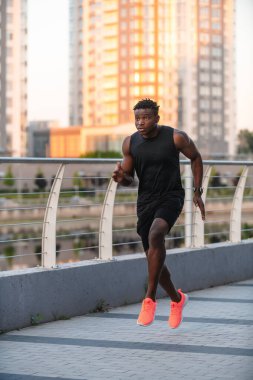 Spor giyimli kendine güvenen Afrikalı adam, arka planda şehir manzarasıyla sabah koşusunun keyfini çıkarıyor.