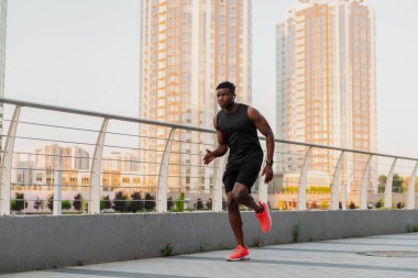 Spor giyim sektöründe kendine güveni olan Afrikalı bir adam arka planda şehir silüeti ile dışarıda koşuyor.