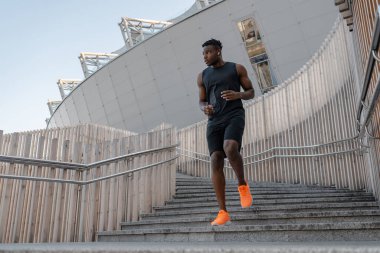 Spor kıyafetleri içinde kendinden emin Afrikalı bir adam açık havada antrenman yaparken merdivenlerden aşağı koşuyor.