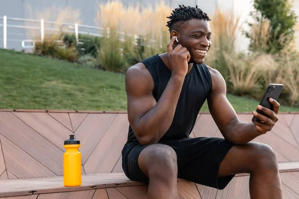 Happy African man in sportswear using smart phone and adjusting headphones while resting after training