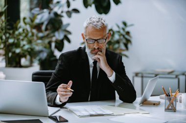 Olgun iş adamı modern ofiste belgelerle çalışırken düşünceli görünüyor.