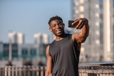 Neşeli genç Afrikalı atlet akıllı telefon kullanıyor ve dışarıyı ararken gülümsüyor.