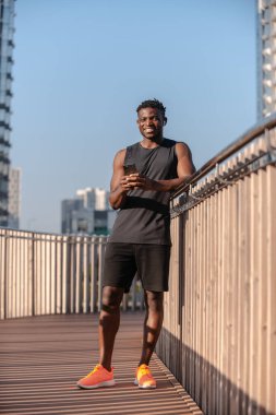Spor kıyafetli yakışıklı Afrikalı adam akıllı bir telefon tutuyor ve dışarıda dururken gülümsüyor.
