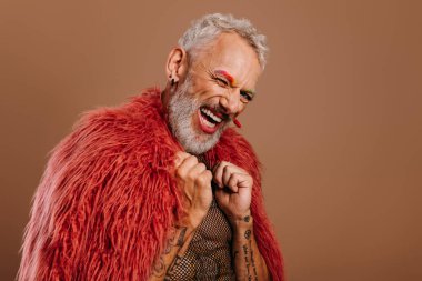 Gökkuşağı renkli kaşları olan, kahverengi arka planda kabarık ceket giyen neşeli, olgun bir gay.