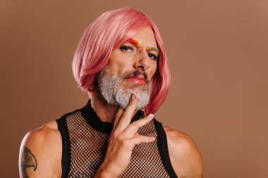 Gökkuşağı renkli kaşları olan, kahverengi arka planda pembe peruk takan olgun eşcinsel bir adamın portresi.