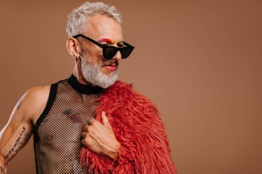 Tarz sahibi, renkli kaşları olan, kahverengi arka planda kabarık bir palto taşıyan, olgun bir gay.