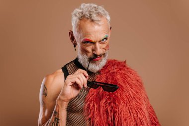 Renkli kaşları olan, kahverengi arka planda gözlük tutan şık, olgun bir gay.