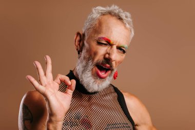 Gökkuşağı renkli kaşları olan neşeli, olgun bir gay. Kahverengi arka planda hareketler yapıp göz kırpıyor.