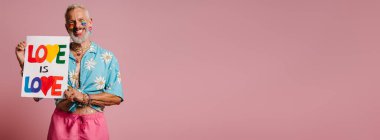 Gökkuşağı bayraklı mutlu, olgun bir gay. Elinde pembe arka planda renkli bir pankart var.