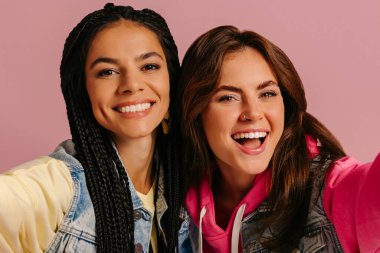 Renkli giyinmiş iki neşeli genç kadın sarılıyor ve pembe arka planda kendi portresini yapıyorlar.