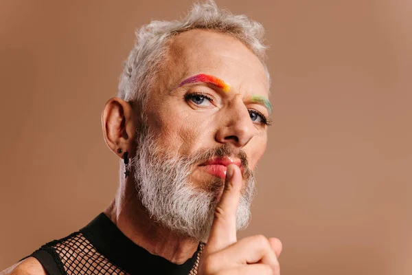 茶色の背景に対して唇に指を保持する虹色の眉毛を持つひげ付き成熟したゲイ男性 — ストック写真