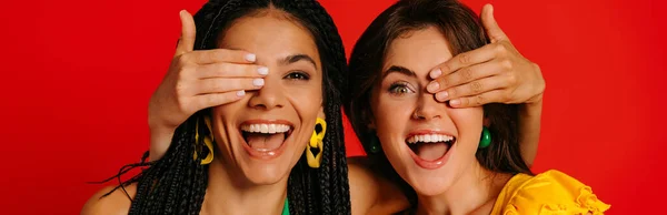 カラフルな身に着けている2人の幸せな若い女性はお互いに目を覆い 赤い背景に微笑みます — ストック写真