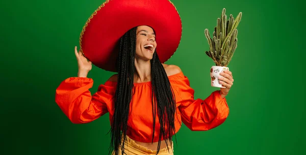 Beautiful Mexican woman in Sombrero holding cactus and looking at it with smile against green background
