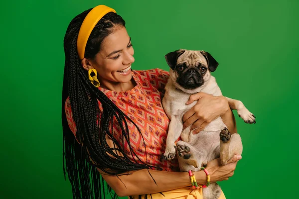 魅力的な若いヒスパニックの女性はかわいいパグ犬を運んで緑の背景に微笑みます — ストック写真