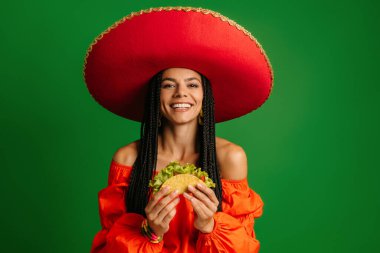 Sombrero 'da elinde taze yapılmış taco ve yeşil arka planda gülümseyen neşeli genç Meksikalı kadın.