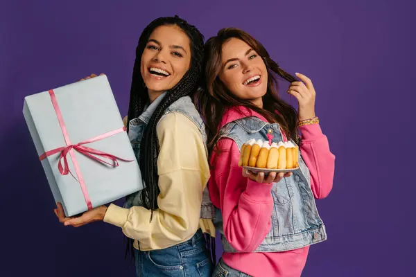 紫色の背景に対して誕生日ケーキとギフトボックスを持っている2人の幸せな若い女性 — ストック写真