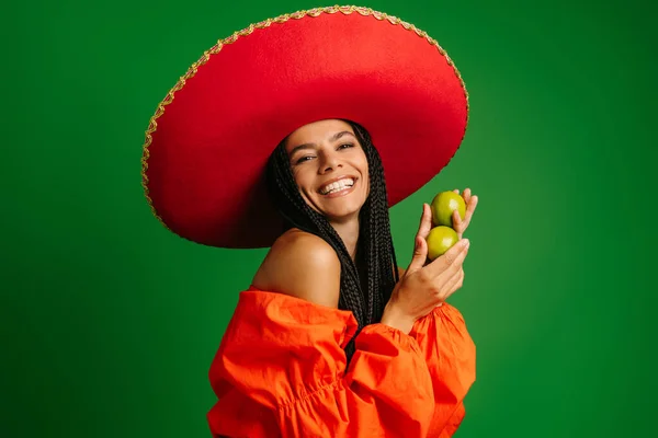 Happy young Mexican woman in Sombrero holding fresh limes and smiling against green background