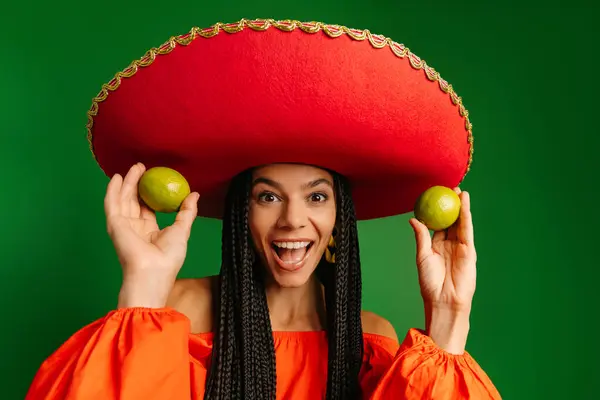 Joyful young Mexican woman in Sombrero showing fresh limes and smiling against green background