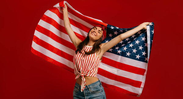 Joyful young hipster woman carrying American flag while standing on red background