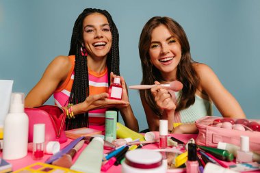 İki mutlu bayan sunucu masasında mavi arka planda otururken çeşitli güzellik ürünleri gösteriyor.