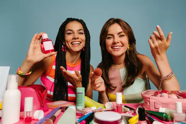 Two happy female presenters showing various beauty products while sitting at the desk on blue background