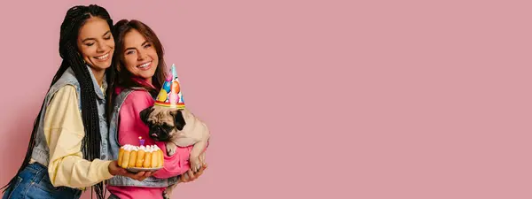 stock image Two joyful young women bonding while carrying cute pug dog and birthday cake against pink background