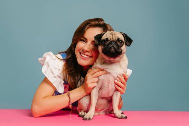 Mavi arka planda pembe masada otururken sevimli pug köpeğine sarılan mutlu genç kadın.