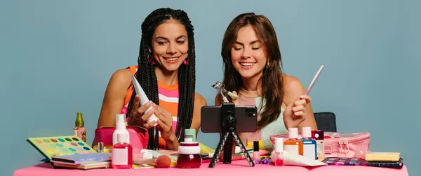 ブルーバックグラウンドでオンラインで放送しながら美容製品を調べる2人の幸せな女性インフルエンサー — ストック写真