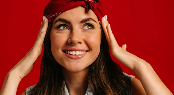 Attractive young hipster woman with colorful make-up touching face and smiling on red background