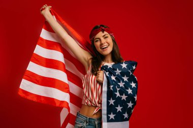 Çekici genç hippi kadın kırmızı arka planda dururken Amerikan bayrağı taşıyor ve gülümsüyor.