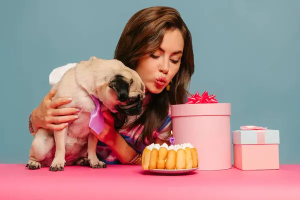 Young woman petting dog and blowing candle on birthday cake while sitting at the pink desk on blue background