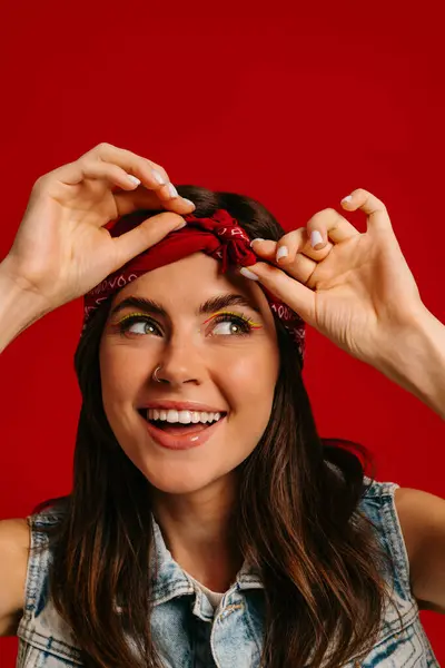 Attractive young hipster woman with colorful make-up adjusting bandana and smiling on red background