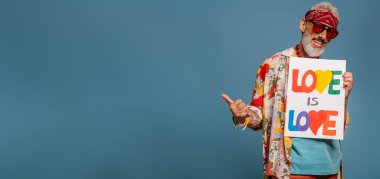 Komik tişörtlü neşeli hippi kıdemli adam elinde afiş ve mavi arka planda el kol hareketi yapıyor.