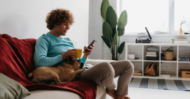 Yakışıklı kıvırcık adam küçük köpeğiyle kanepede dinlenirken akıllı telefon kullanıyor.