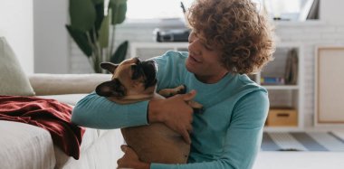 Yakışıklı kıvırcık adam sevimli köpeğini tutuyor ve evde dinlenirken gülümsüyor.