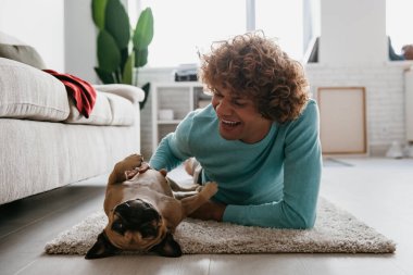 Mutlu kıvırcık adam evde yerde birlikte yatarken sevimli köpeğini okşuyor.