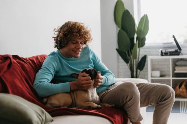 Kulaklıklı yakışıklı kıvırcık adam evdeki kanepede dinlenirken sevimli köpeği okşuyor.