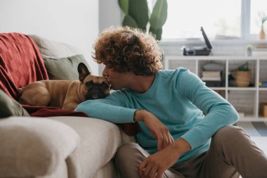 Yakışıklı kıvırcık adam evde dinlenirken tatlı köpeğini öpüyor.