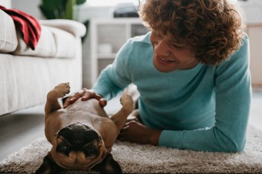 Neşeli genç kıvırcık adam evde yerde birlikte yatarken sevimli köpeğini okşuyor.