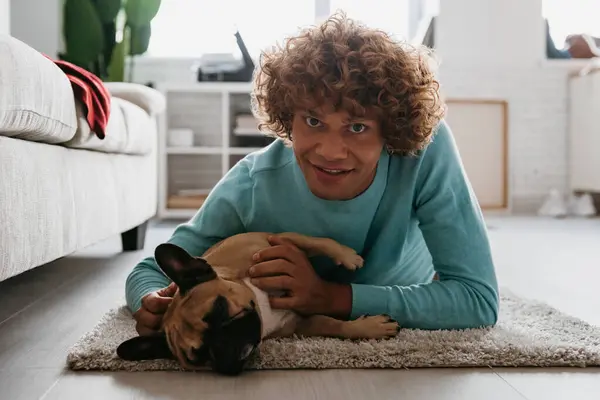 Handsome young curly man petting his cute dog and smiling while lying on the floor at home together