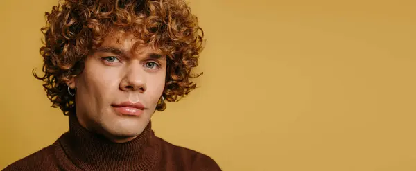 Handsome young curly man in sweater looking at camera against yellow background