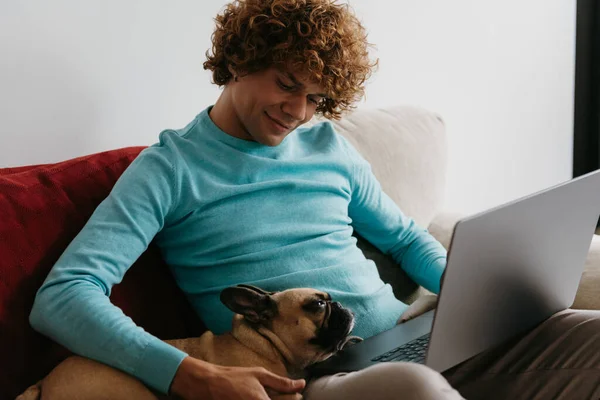 Handsome young curly man petting his dog while working on laptop from home