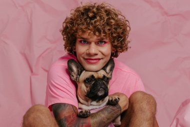Yakışıklı genç eşcinsel adam sevimli köpeğini tutuyor ve buruşuk pembe arka planda otururken gülümsüyor.