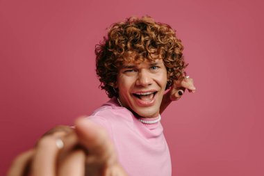 Oyuncu kıvırcık saçlı adam pembe arka planda kameraya gülümsüyor.